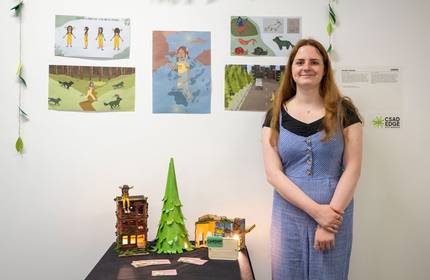 A woman in a blue dress stands next to a display table with a small model of a building and a tree. The wall behind her has colourful illustrations and a CSAD edge logo.