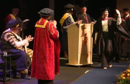 A young woman in cap and gown walks across a stage during a graduation ceremony.