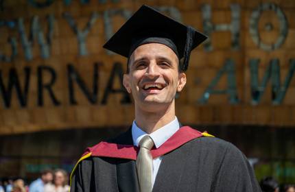 A man wearing graduation robes and a cap, standing in front of Wales Millennium Centre.