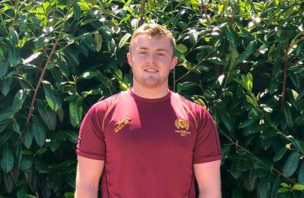 A person wearing a maroon sports jersey smiles while standing in front of a lush green hedge.
