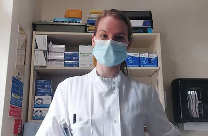 A healthcare professional wearing a white coat and a blue surgical mask stands in front of shelves stocked with medical supplies and gloves. The setting appears to be a clinic or a hospital.