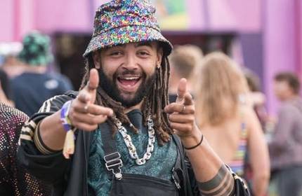 A man with dreadlocks and a colourful bucket hat smiles and points finger guns at the camera. He wears a turquoise patterned shirt, layered necklaces, and stands in a crowd at an outdoor event. The background is pink and blurred.