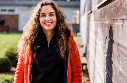 A person with long curly hair smiles while standing outside. They are wearing a red jacket over a black top. The background features a wooden wall and grass, with a softly blurred building in the distance.