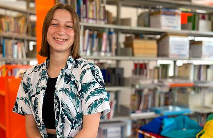 A smiling person stands in a library wearing a tropical-patterned shirt over a black top. Shelves filled with books and colourful bins are visible in the background, creating a vibrant and welcoming atmosphere.