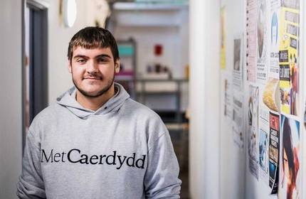 A person with short hair and a beard wearing a gray Met Caerdydd hoodie stands in a hallway with various posters on the wall. The background features a corridor with soft lighting.