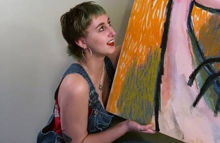 A person with short hair and hoop earrings, wearing overalls and a floral top, crouches while holding a colourful abstract painting. They look upwards with a smile, set against a neutral background.