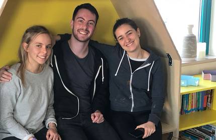 Three people sitting closely in a cozy corner with a yellow background, smiling at the camera. Two women are on either side of a man, each wearing casual clothing. A bookshelf with colourful items is partially visible in the corner.