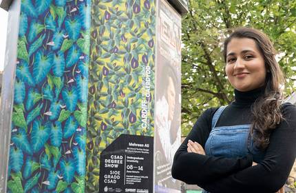 A woman stands confidently with crossed arms in front of a vibrant art installation featuring colourful leaf patterns. Shes wearing a black turtleneck and denim overalls. A tree and part of a building are visible in the background.