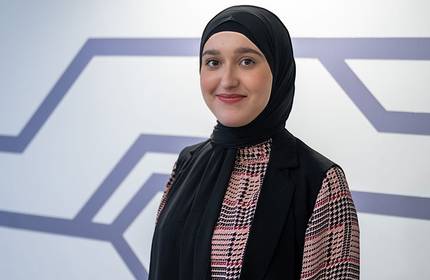 A person wearing a black hijab and a plaid shirt smiles softly while standing in front of a geometric wall design.