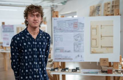 A young man stands in front of a display of architectural drawings and small models of buildings.