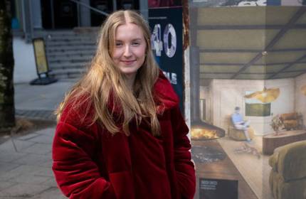 A young woman in a red jacket stands next to an outdoor poster display. One of the posters is an illustration of a living room.