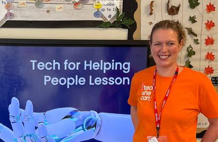 A woman wearing an orange t-shirt stands in front of a large screen. A caption on the screen reads 'Tech for Helping People Lesson'.