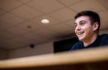 A smiling person sits in a lecture room.