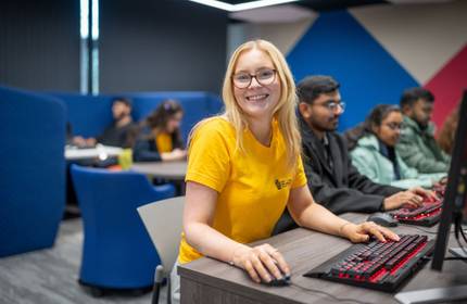 A young woman using a computer workstation types on an keyboard with illuminated red keys. In the background are other people using similar workstations.