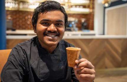 A young man holds a small edible coffee cup made from biscuit. Behind him is a bar area with a coffee machine.