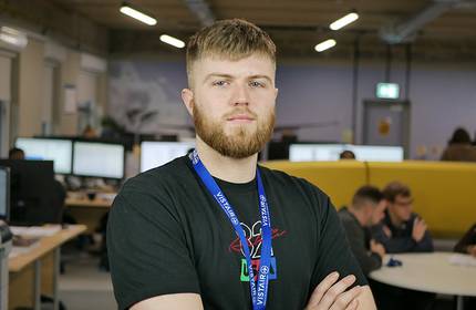 A man with a beard stands confidently in an office environment wearing a blue lanyard. Behind him, several people are working at desks with computer monitors. The office has an industrial design with exposed ceilings.