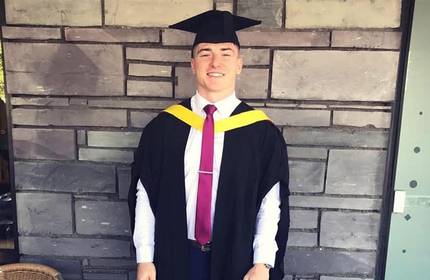 A man wearing academic robes over a shirt and tie stands in front of a wall.
