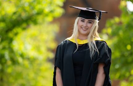 A young woman wearing a graduation cap and gown stands smiling. In the background is green foliage.