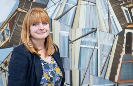 A woman with red hair stands in front of an abstract, geometric painting featuring overlapping architectural elements. She wears a patterned top under a dark jacket and smiles gently at the camera.