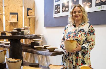 A person holds a carved bowl. A number of similar bowls on wooden stands surround them.