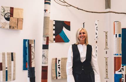 A woman stands in front of a display of patterned fabrics. Some are mounted on the wall, while others are wrapped around poles.