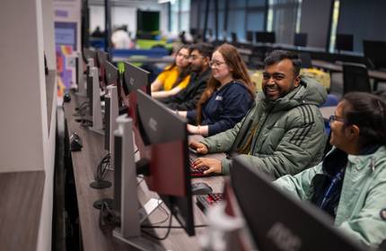 A group of five students sit alongside each other at a long table. Each is using a computer workstation.