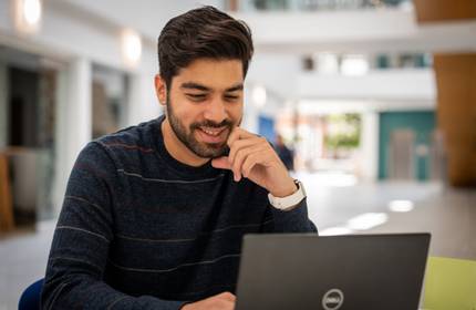 A young man sits in front of a laptop in a bright atrium space.