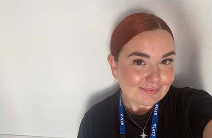 A person with red hair is smiling at the camera, wearing a black shirt and a blue lanyard with text. The background is plain and light-coloured.