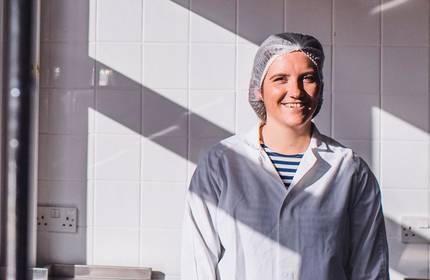 A person wearing a white lab coat and a hairnet stands smiling in a brightly lit room with white tile walls. Shadows from window panes fall across the wall, creating a pattern of light and shadow.