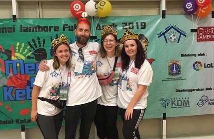 Four people wearing matching white T-shirts and traditional woven hats stand smiling in front of a colourful banner. The banner features logos and text related to a football event. Balloons are visible at the top.
