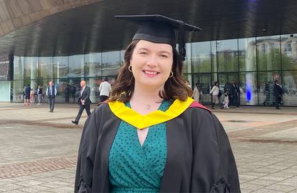 A person in a graduation cap and gown smiles outdoors. They are standing in front of a modern glass building, with several people walking in the background.