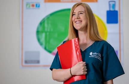 A person with long red hair, wearing teal scrubs, smiles while holding a red folder. The background features a colourful wall chart with various symbols and text.