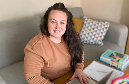 A person with long hair and a brown sweatshirt sits at a desk with a laptop, smiling at the camera. The background features a grey sofa with patterned pillows and a stack of colourful books.