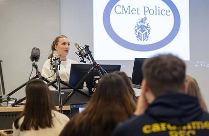 A woman is speaking at a podium with several microphones in front of her. A logo for CMet Police is displayed on a screen behind her. Several people are seated, facing the speaker.