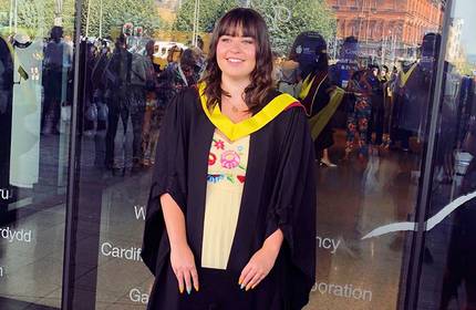 A person in a graduation gown and cap smiles while standing outside a glass-fronted building. They wear a white dress with colourful embroidery underneath their gown. Reflections of people and trees are visible in the glass.