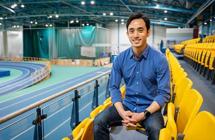 A young man sits in an empty row of seats inside an indoor athletics facility. A running track is visible in the background.