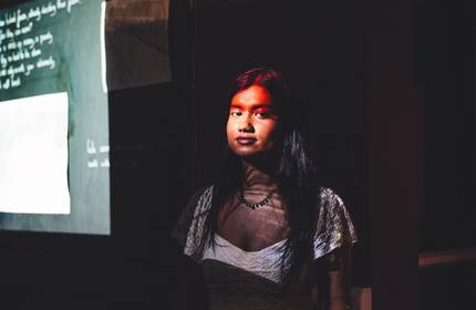 A young woman stands in a darkened room, her face illuminated with red light.