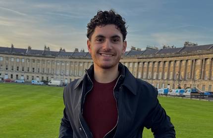 A young man in black jacket smiles while photographed on an open field in front of a row of tall houses