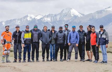 A large group of people stand in a row. In the background is a mountain range.