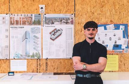 A young man wearing a black t-shirt stands with his arms folded in front of wooden boards. Mounted on the boards are plans and designs for a tower block.