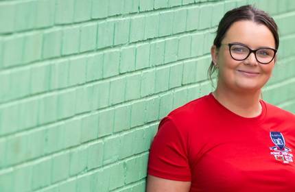 A smiling person leans against a green painted brick wall.