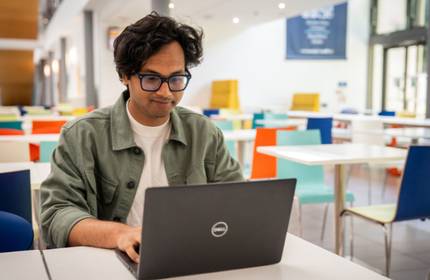A young man sits at a table working on a laptop.