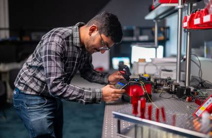 A student pressing the touchscreen of a fibre optic splicer. In the background are other devices and computer monitors.