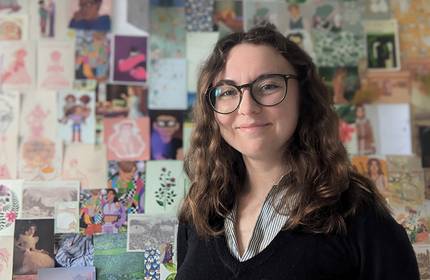 A person stands smiling in front of a wall covered with postcards and prints.