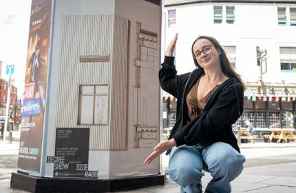 A young woman next to an outdoor advertising display. The display includes a poster showing an architectural drawing and the text DEGREE SHOW.