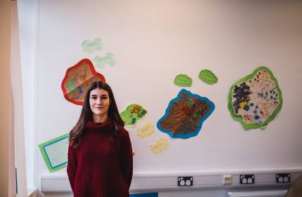 A person with long dark hair wearing a red sweater stands in a classroom setting. The background shows colourful artwork displayed on the wall.