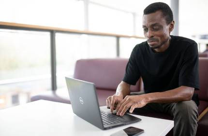 A person sits at a table typing on a laptop computer. There is a mobile phone next to the laptop.