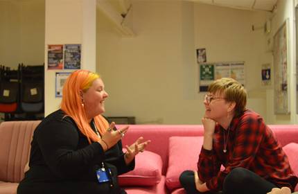 Two smiling people sit on a pink couch. One has long, orange hair and gestures while holding a phone. The other, wearing glasses and a red plaid shirt, leans forward with a hand under their chin, engaged in conversation. Posters and chairs are in the background.