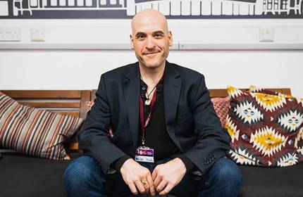 A bald man wearing a black blazer is sitting on a dark sofa. He is smiling and has his hands clasped. The sofa has various patterned cushions. The background features a black and white wall design.