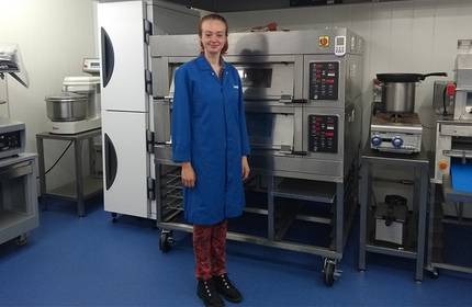 A person in a blue lab coat stands in a commercial kitchen next to industrial ovens and equipment. The room has a blue floor and stainless steel appliances.